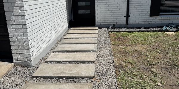 Concrete stepping stones lead to a black door beside a white brick wall.
