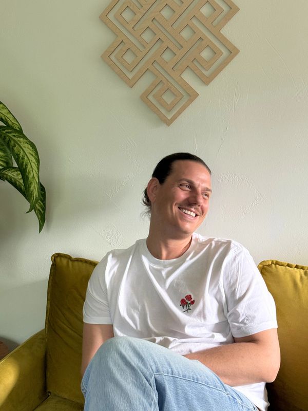 Smiling man sitting on a mustard yellow couch, wearing a white t-shirt with a rose design.