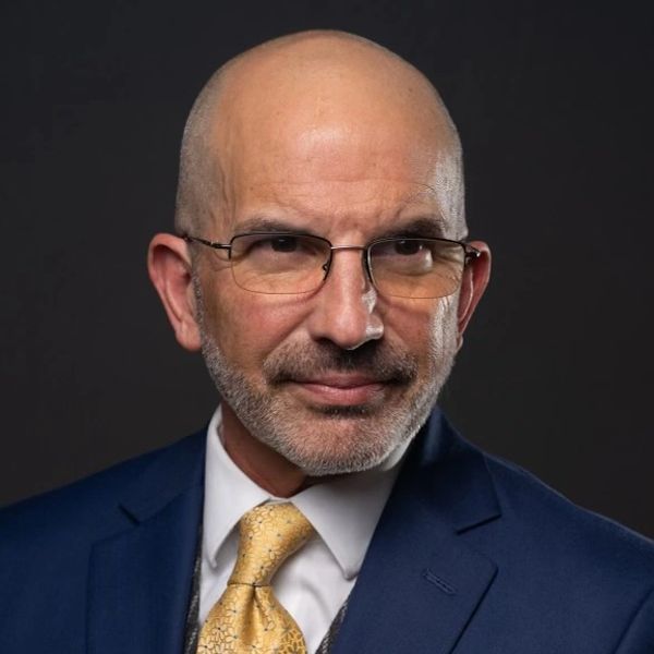 Confident bald man in a blue suit with glasses and a gold tie.