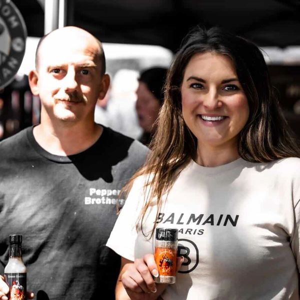 Two people smiling, holding hot sauce bottles, one wearing a Pepper Brothers shirt.