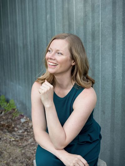 Smiling woman in a teal dress sitting outdoors against a corrugated metal wall.