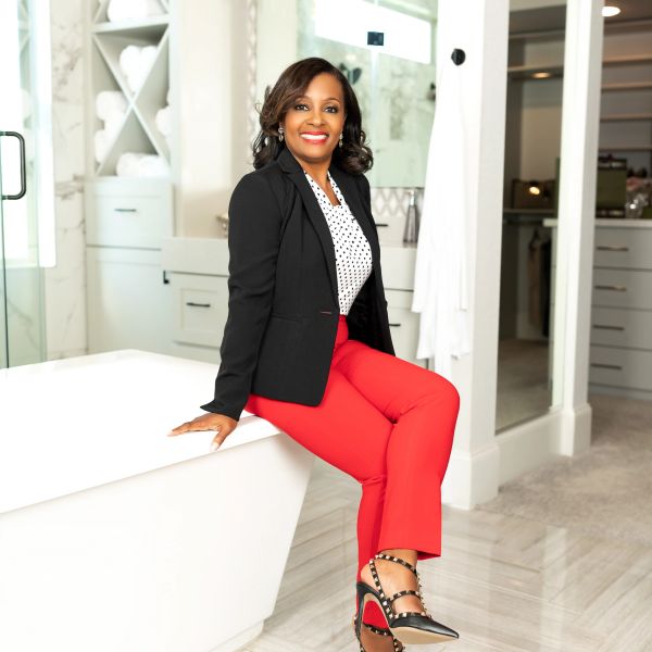 Confident woman in red pants and black blazer sitting on a bathtub edge.