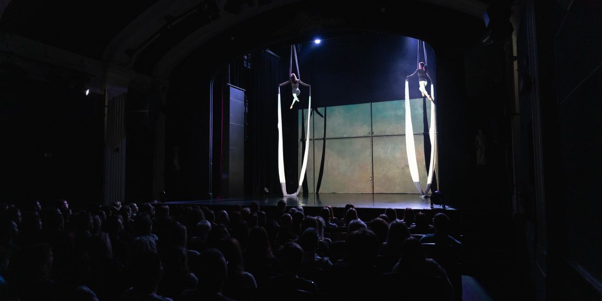 Two aerial silk performers on stage in front of a seated audience.