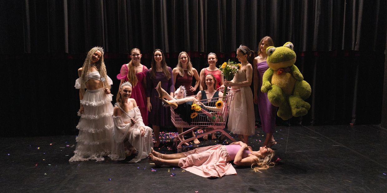 A group of women in elegant dresses posing with a shopping cart and large frog plush on stage.