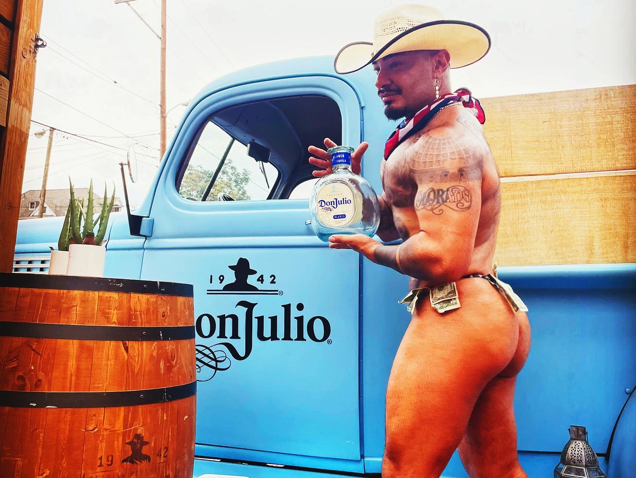 Muscular man in cowboy hat holding Don Julio tequila bottle by blue truck.