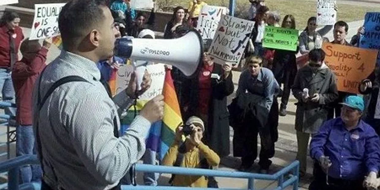 Man speaks through megaphone at equality rally with diverse crowd holding signs.