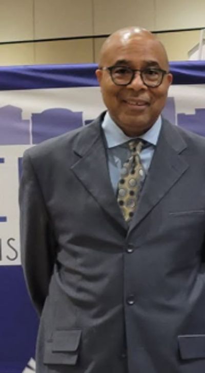 Man smiling, wearing glasses and a suit with a patterned tie.