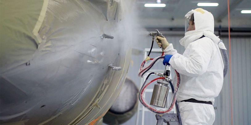 Worker in protective suit spray painting an aircraft fuselage indoors.