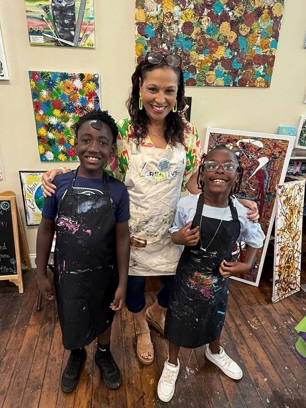 Three people smiling in a colorful art studio, wearing paint-splattered aprons.