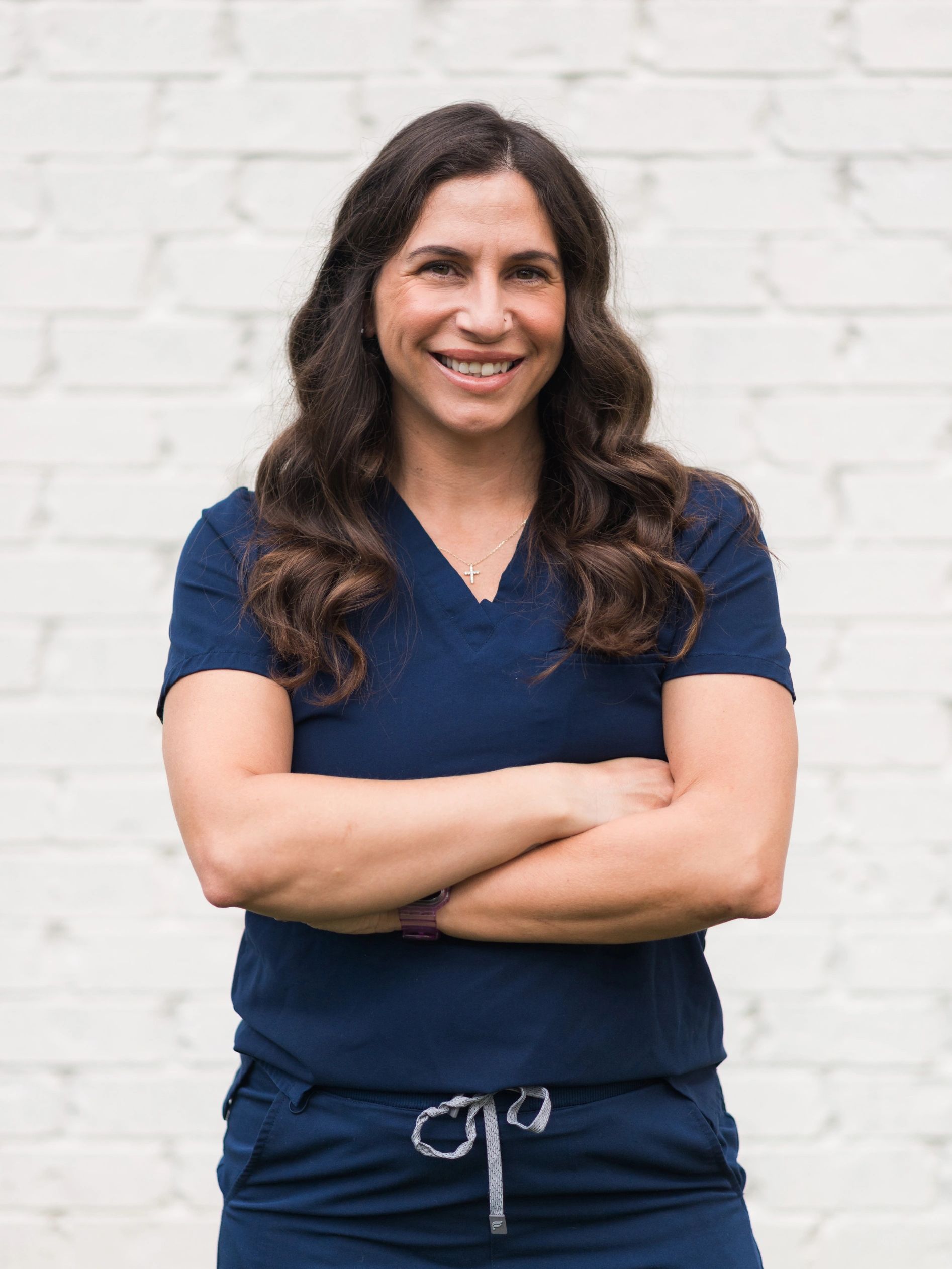 Smiling woman in navy blue scrubs with arms crossed, standing against a white brick wall.
