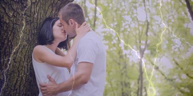 Two young adults kissing against a tree with lightning in the background.