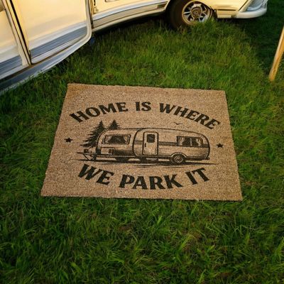 A doormat with a camper and the phrase 'Home is where we park it' on grass.
