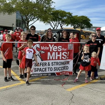 Group of people holding a banner supporting voting Yes for MHS in a parking lot.