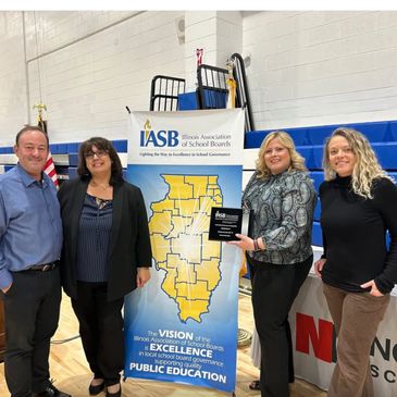 Four people stand beside an Illinois Association of School Boards banner holding an award plaque.
