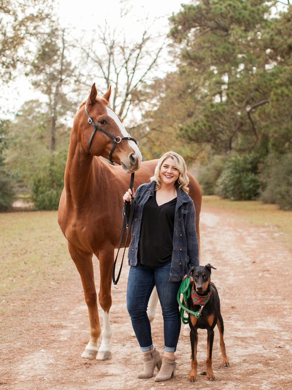 Woman standing with a horse and a dog on a dirt path.