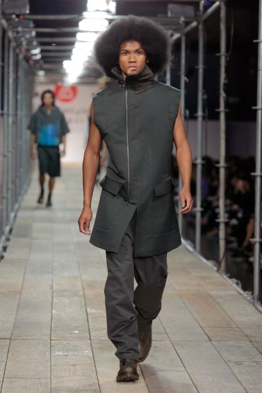 Male model with afro hairstyle walking on runway in sleeveless dark outfit.