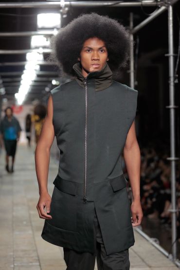 Male model with afro hairstyle in sleeveless dark green vest on runway.