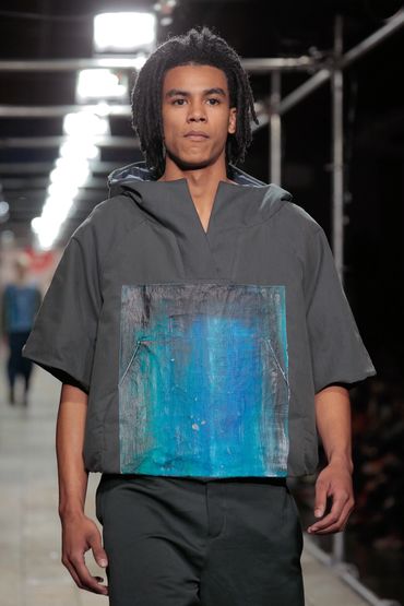 Male model wearing a gray top with a blue artistic panel on runway.