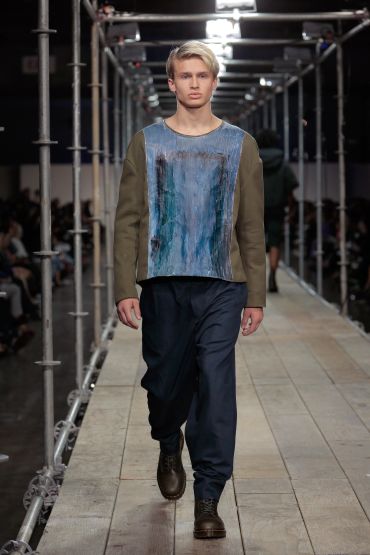Model walking down a runway in a blue and olive sweatshirt with dark pants and boots.