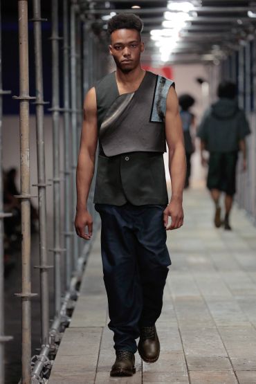 Male model walking runway in asymmetrical black sleeveless jacket and dark pants.