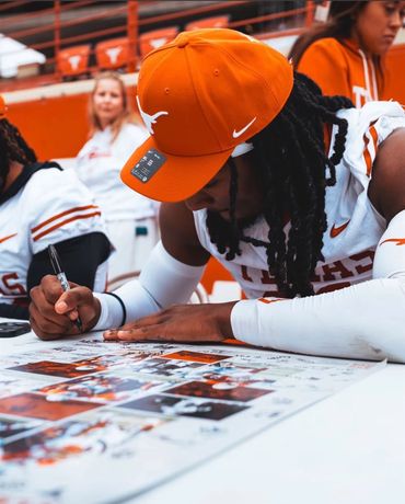 A Texas Longhorns football player signs a poster while wearing an orange cap.