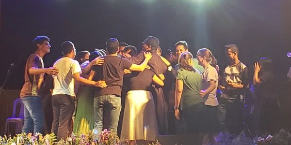 A group hug on stage with diverse people under bright lights.