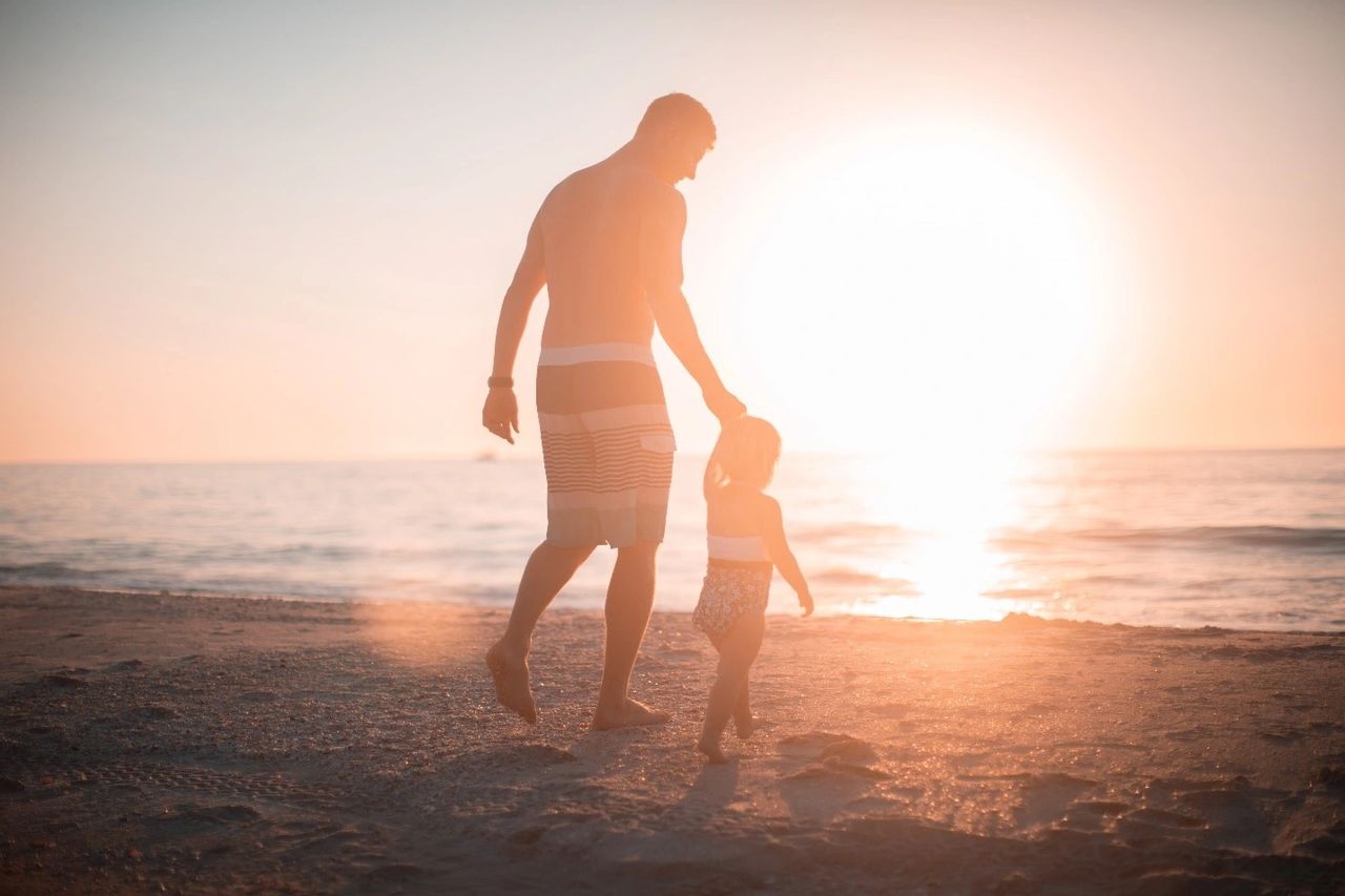 A father holding his child’s hand at the beach.