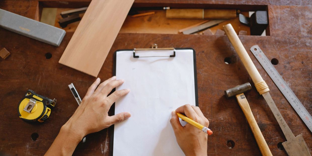 Hands sketching woodworking plans surrounded by tools on a wooden table.