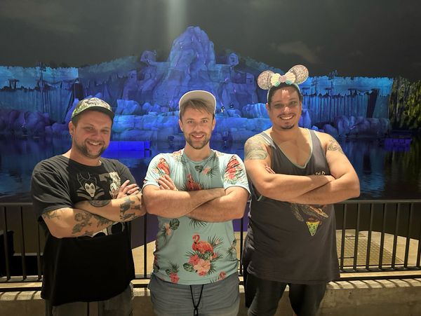Three men posing with arms crossed in front of a lit water and rock backdrop at night.