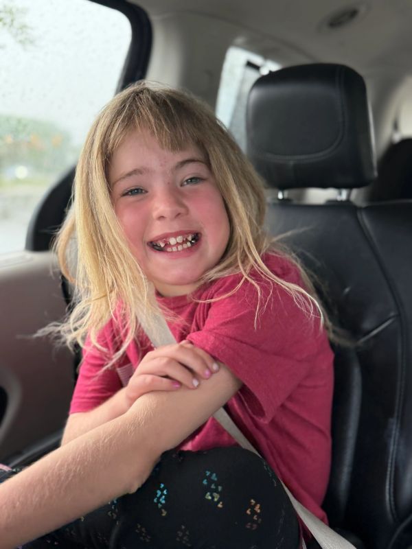 Smiling young girl with blonde hair sitting in a car seat, wearing a red shirt.
