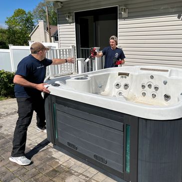 Two men moving a large hot tub outside a house on a sunny day.