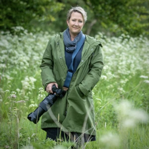 Woman in green coat holding a camera in a field of greenery.