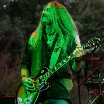 Long-haired guitarist playing electric guitar on stage with green lighting.