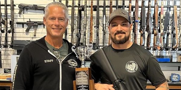 Two men posing with a trophy and rifles in a gun store.
