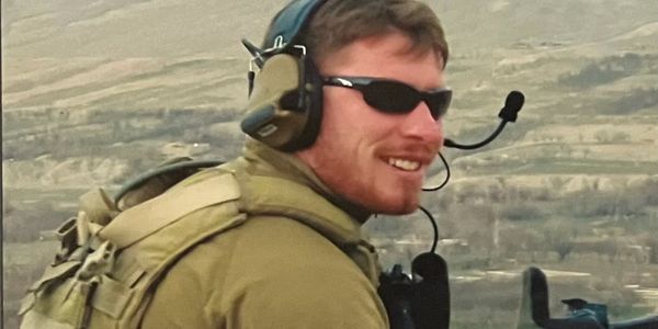 Soldier in tactical gear with headset and sunglasses smiling in a mountainous area.