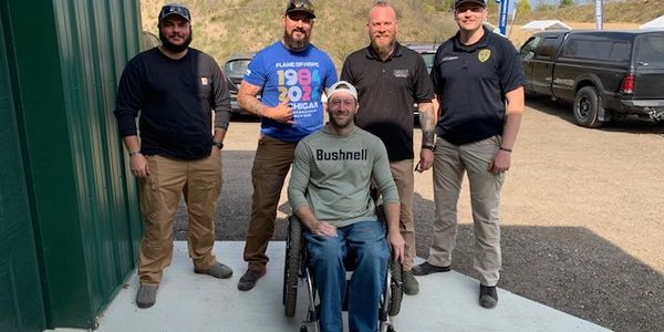 Five men posing together outdoors, one seated in a wheelchair.