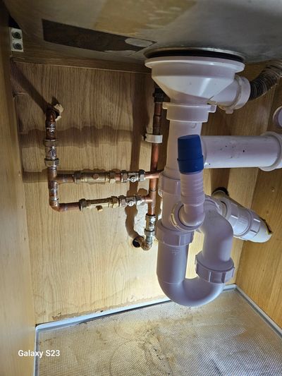 Plumbing under a sink with copper and white pipes inside a wooden cabinet.