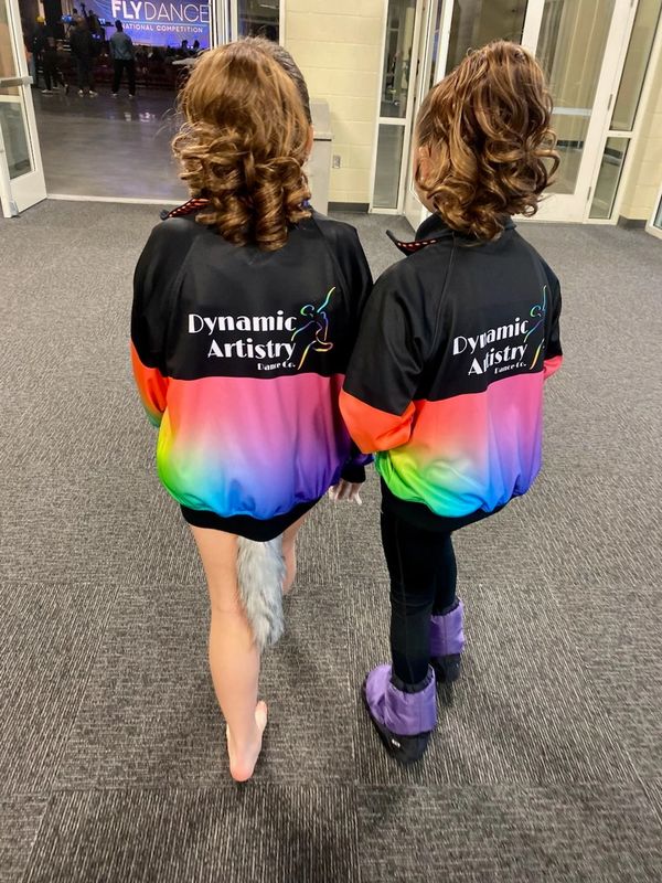 Two dancers in colorful Dynamic Artistry jackets at a dance competition venue.