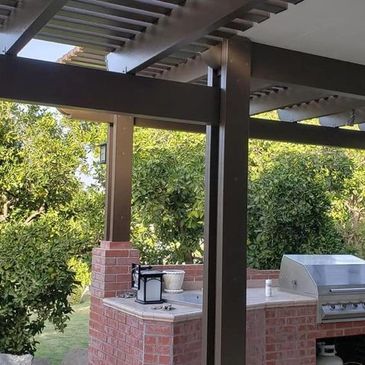 Outdoor brick barbecue grill setup under a pergola with green trees in the background.