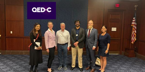 Group photo of six professionals standing indoors near a QED-C logo.