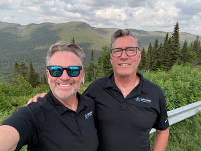 Deux hommes souriants en polo noir en pleine nature.