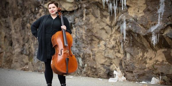 Woman posing with cello outdoors near rocky wall with icicles.