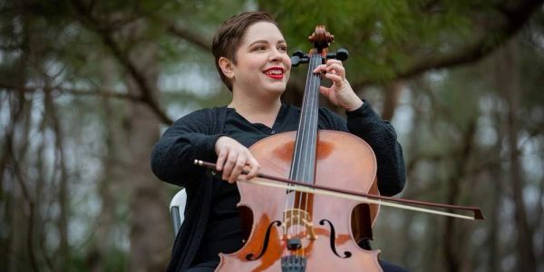 Person playing cello outdoors, smiling.