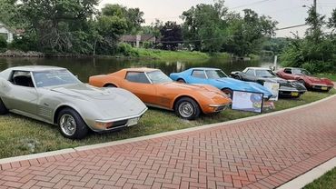 Five classic cars parked near a river on grass beside a brick walkway.