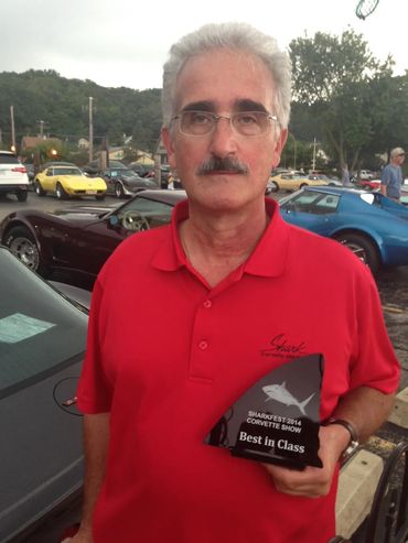 Man in red shirt holding Best in Class award at a car show.