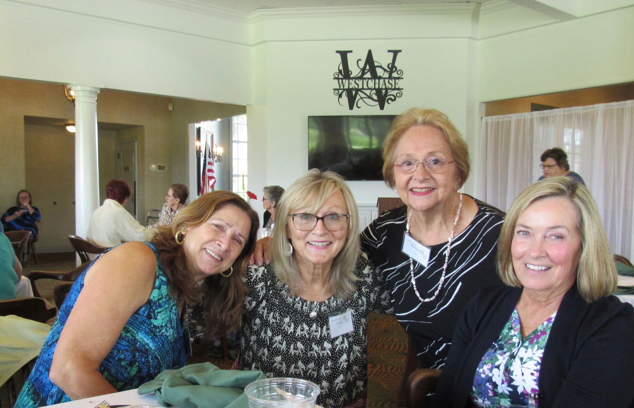 Four smiling women at a social gathering inside a room with a Westchase sign.