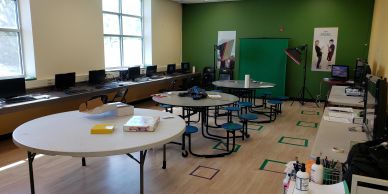 A modern classroom with round tables, computers, and a green screen area.