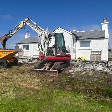 Excavator working on rocky ground near a white house under a clear blue sky.