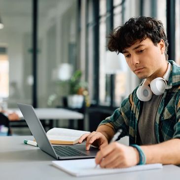 boy with headphone around his neck studying from laptop and making notes
