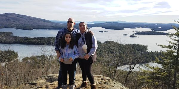 A family of three stands on a rock with a scenic lake and mountains in the background.